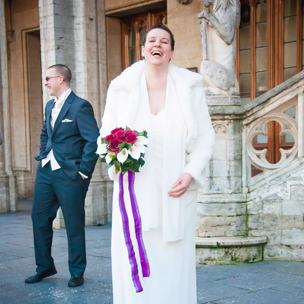 Mariée riant avec son bouquet devant un bâtiment en pierre, le marié à ses côtés